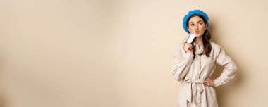 Shopping and discounts concept. Young stylish modern girl, posing with credit card, looking happy and smiling, standing over beige background.