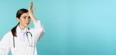 Portrait of annoyed, tired woman doctor, facepalm, roll eyes frustrated, bothered by smth stupid, standing in white coat over torquoise background.