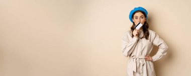 Shopping and discounts concept. Young stylish modern girl, posing with credit card, looking happy and smiling, standing over beige background.