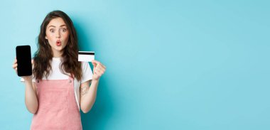 Plastik kredi kartı ve boş akıllı telefon ekranı gösteren genç güzel bir kadının portresi, eğlenceli ve ilgili görün, ilginç bir uygulama gösteriyor, mavi arka planda duruyor..