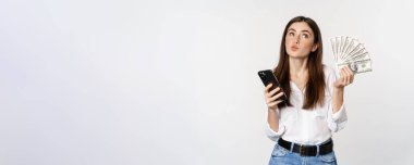 Portrait of happy smiling woman using mobile phone app, holding money cash, standing over white background. Copy space