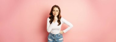 Glamour stylish brunette girl, shush, silence you, press finger to lips and winking, sharing secret, hushing, be quiet sign, standing over pink background.
