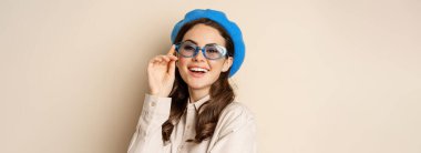 Close up portrait of stylish woman posing in sunglasses, laughing and smiling, wearing trendy outfit, standing over beige background.