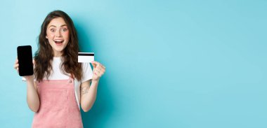 Bana bak. Boş cep telefonu ekranı ve plastik kredi kartı gösteren heyecanlı genç kız mavi arka planda dikilen kameraya hayret içinde bak..