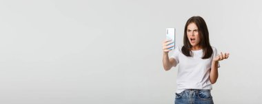 Hüsrana uğramış genç bir kadın video görüşmesinde tartışıyor, elinde akıllı telefon, ayakta duran beyaz arka plan..