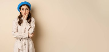 Image of stylsh beautiful woman looking surprised, shocked reaction at camera, posing in trench coat against beige background.