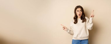 Confused girl shopper pointing sideways, making choice, decision between two variants, beige background. Copy space