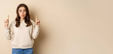 Surprised beautiful girl showing advertisement, pointing fingers on top, store sale or website link up, standing over beige background.