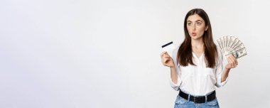 Excited woman holding credit card and money, looking amazed at cash, standing against white background. Copy space