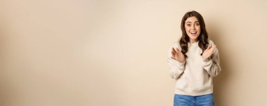 Image of surprised, amazed brunette girl gasping, looking fascinated at camera, standing over beige background.