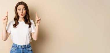 Excited girl looking interested, pointing fingers up, showing advertisement, standing in tshirt over beige background.