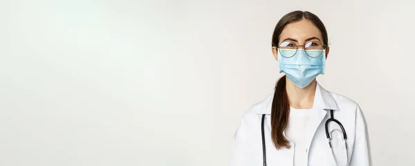 stock image Portrait of medical worker, female physician in face mask from covid during pandemic, smiling and looking enthusiastic, standing over white background.