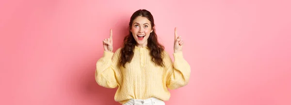 stock image Excited brunette girl pointing fingers and looking up with excited face, reading promotional text, checking out advertisement, standing against pink background.