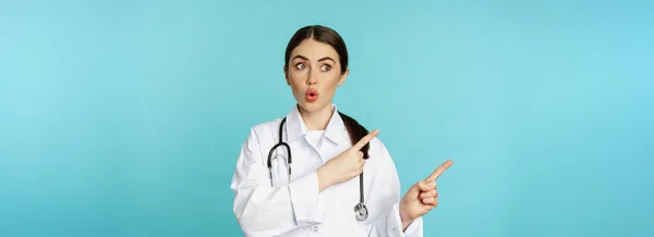 驚くべき女性医師 聴診器を持つ医師 指差し 驚くべき すごい顔で左を見て ターコイズの背景の上に立って — ストック写真