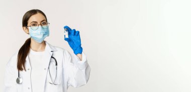 Genç kadın portresi, tıbbi maske ve üniformalı doktor, aşıyı gösteriyor, covid-19 aşı kampanyası, beyaz arka planda duruyor..