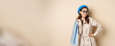 Stylish happy girl on shopping. Portrait of modern woman with shop bag, laughing and smiling satisfied, buying herself gift, standing over beige background.