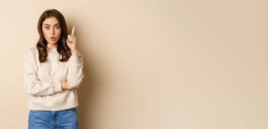 Young woman suggests an idea, raising finger, has revelation, pitching a plan, pointing up, standing over beige background.