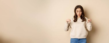 Surprised girl pointing and looking down at smth curious below, beige background.