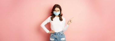 Angry young woman in medical face mask, frowning frustrated, pointing finger left, showing promo copy space, standing over pink background.