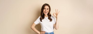 Beautiful female model smiling pleased, showing okay, zero sign, complimenting, praising you, standing over beige background.
