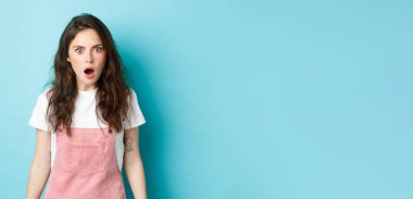 Omg ne? Şok olmuş, kıvırcık saçlı, çenesi düşük, nefes nefese, şok edici bir şeye inanmayan, büyük haberleri duyan, mavi arka planda duran genç kız..