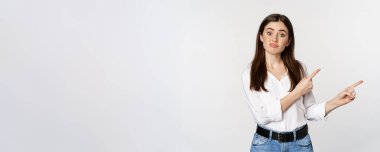 Sad and disappointed cute woman, gloomy girl pointing fingers at logo, complaining, sulking upset, standing in blouse over white background.