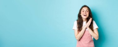 Mutlu bir kadının portresi, iyi haberlerle seviniyor, el çırpıyor ve gülümsüyor, hediyeye teşekkür ediyor, tatmin olmuş görünüyor, mavi arka plana karşı duruyor..