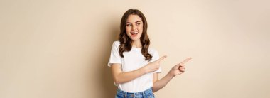 Stylish young caucasian woman smiling, pointing fingers left, showing advertisement, promo offer, standing against beige background.