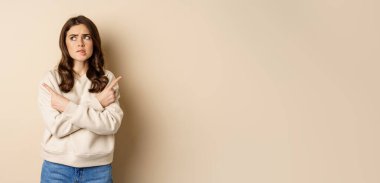 Complicated and indecisive woman pointing sideways, cant make choice, struggle with decision, choosing between two options, standing over beige background.