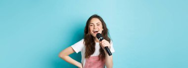Neşeli genç bir kadın karaoke söylüyor, mikrofon tutuyor ve gülümsüyor, eğleniyor, mavi arka planda duruyor..