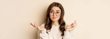 Young indecisive woman in glasses shrugging shoulders and looking confused, dont know, cant understand, standing over beige background.