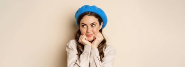 Close up portrait of feminine young woman in trendy french hat, posing cute and romantic, making silly coquettish face and tender smile, beige background.