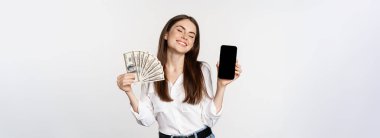 Portrait of smiling woman showing mobile phone screen and money cash, looking happy, standing over white background.