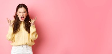 Angry and pissed-off girl screaming, shaking hands and grimacing furious, stare with outraged annoyed face at camera, standing against pink background.