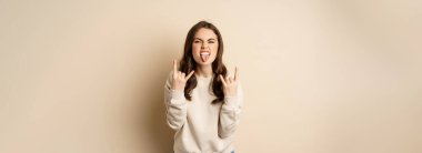 Happy young woman enjoying music, having fun, showing rock on, heavy metal finger horns gesture, standing over beige background.