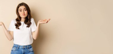 Young confused woman shrugging shoulders and with clueless face expression, standing over beige background.