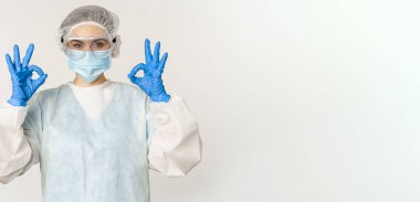 Doctor in personal protective equipment and face mask, showing okay, ok gesture, standing over white background. Healthcare and covid-19 concept