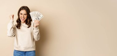 Enthusiastic modern woman winning money, got cash, celebrating and shouting of joy, standing against beige background.