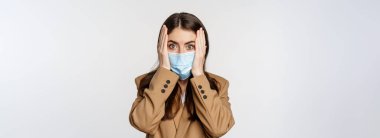 Covid-19 and people concept. Concerned, troubled office lady in medical face mask, holding hands on head and staring shocked at camera, white background.