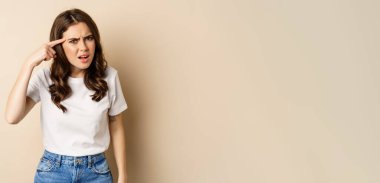 Annoyed and angry woman pointing finger at head, scolding someone, standing over beige background.
