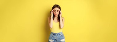 Stylish modern woman rubbing temples, having headache, migraine, frowning and grimacing from pain in head, standing against yellow background.