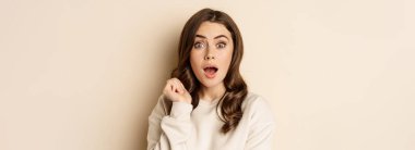 Close up shot of excited woman looking amazed with opened mouth, standing over beige background. Copy space