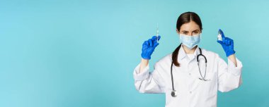 Gülümseyen sağlık personeli, maske takan doktor ve lastik eldiven, mavi arka planda duran covid-19 salgınından şırınga ve aşı gösteriyor..
