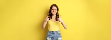 Portrait of stylish modern girl, feminine woman showing thumbs up, recommending gesture, like or approve, praise, standing over yellow background.