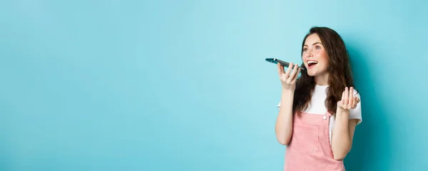 stock image Young woman using app translator on smartphone, speaking into mobile phone dynamic, holding cellphone near lips, recording voice message, blue background.