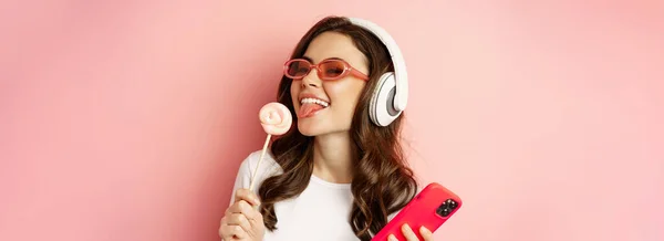 漂亮的女模特戴着耳机听音乐 拿着棒棒糖和手机 戴着太阳镜 站在粉色的背景上 — 图库照片