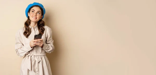 stock image Dreamy smiling woman in stylish trenchcoat, looking up fantasizing while shopping on mobile phone app, using smartphone and thinking, standing over beige background.