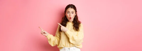 stock image Portrait of surprised brunette girl gasping impressed, pointing fingers right at copy space aside, showing way to logo, standing over pink background.