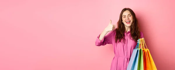 stock image Young happy female shopper showing thumbs up, pleased with good discounts, buying staff on sale, holding shopping bags and smiling pleased, pink background.