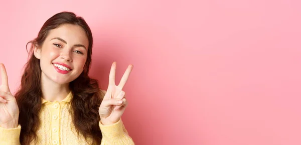 stock image Close up of beautiful young woman showing peace v-sign and smiling happy at camera, wearing bright glam make up, standing against pink background.